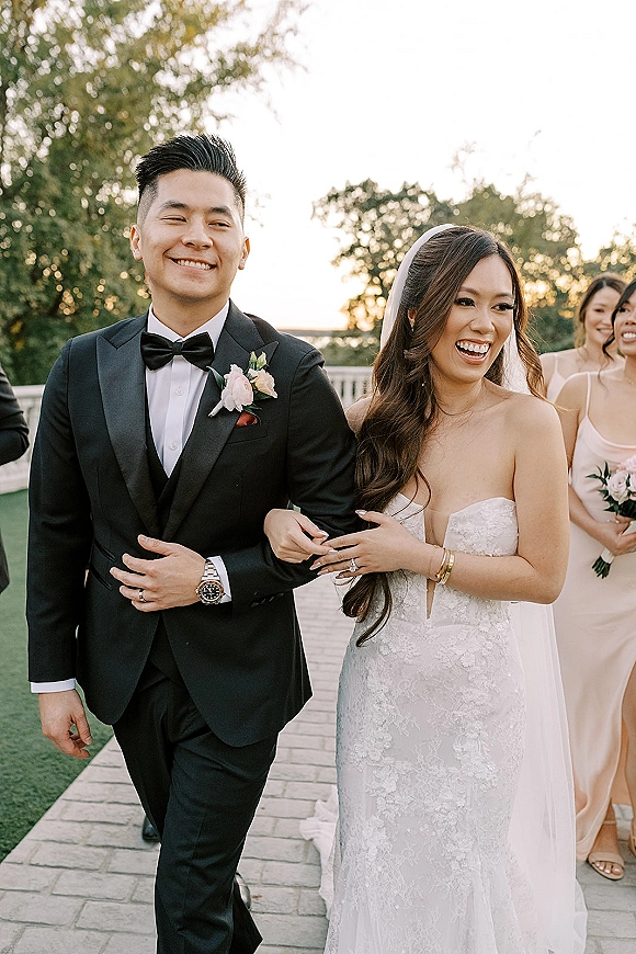 Wedding recessional as bride and groom walking arm in arm, laughing in lace gown and black tuxedo, with bridal party on tree-lined walkway with white railing