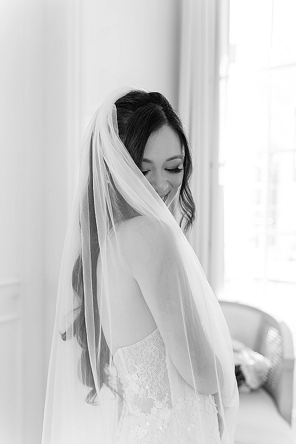 Bridal portrait in black and white of a bride looking down, veil over her face and strapless lace dress in soft window light indoors