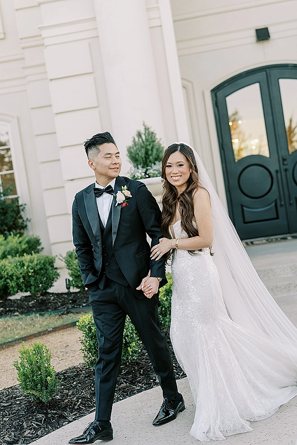 Couple portrait of bride and groom walking arm in arm, her cathedral veil flowing, past black double doors on venue steps