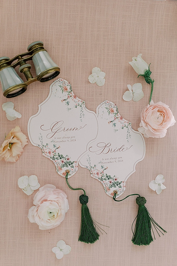 Wedding place cards with bride and groom place cards, floral borders, green tassels, and binoculars arranged on a blush linen backdrop