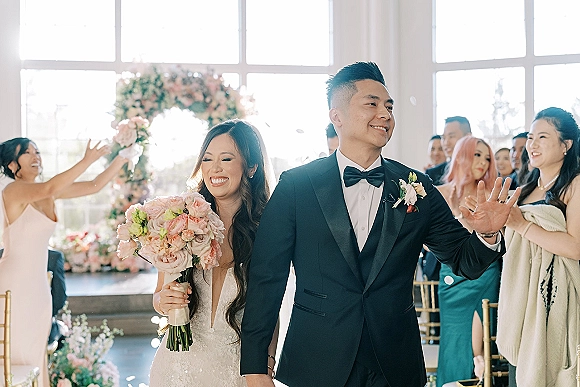 Wedding recessional as newlyweds walking down aisle under window light, bride with bouquet and veil as guests cheer amid confetti petals