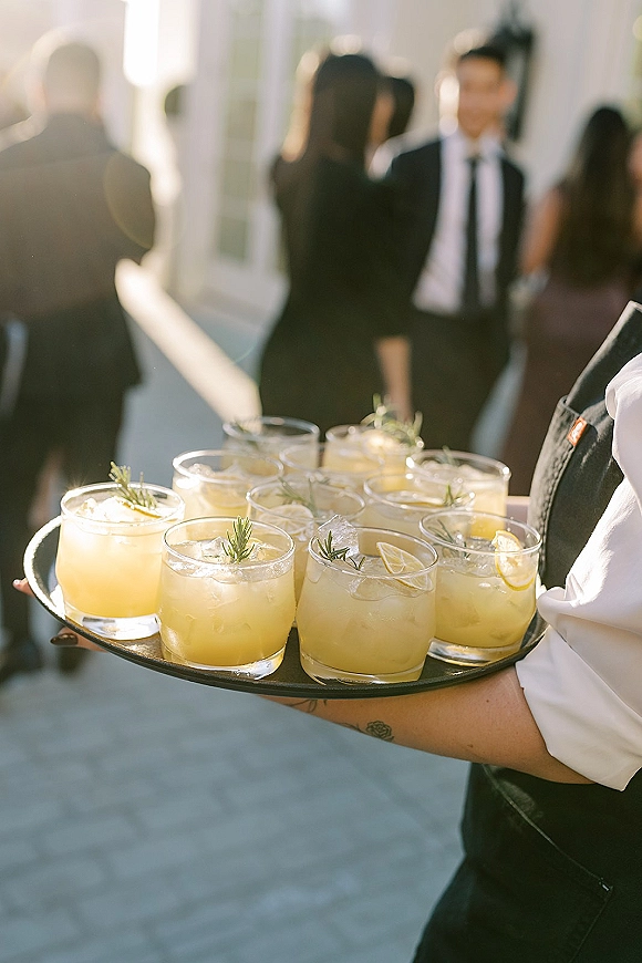 Signature cocktails on a wedding cocktail tray with lemon slices and rosemary sprigs in rocks glasses on a sunlit outdoor patio with guests