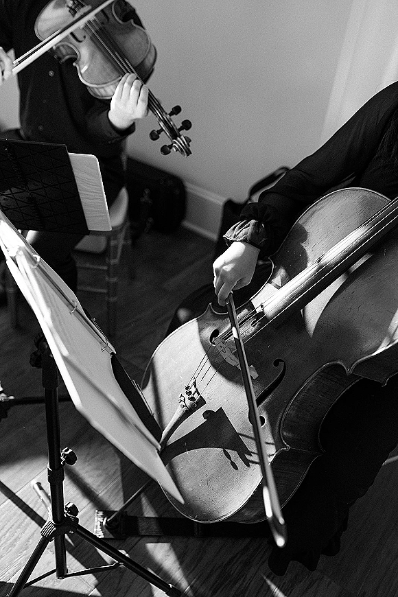Wedding musicians perform as a string duo wedding, cello and violin reading sheet music at stands in black attire by window light indoors