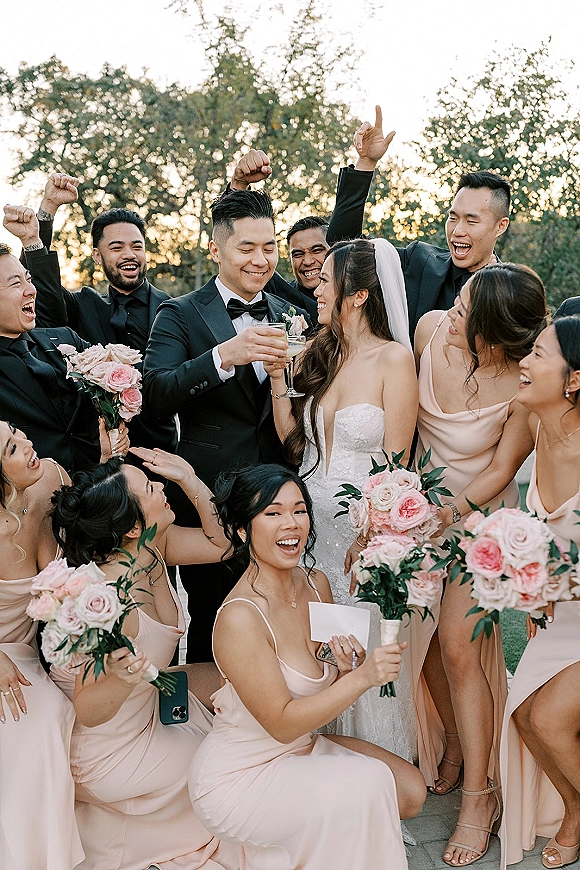 Wedding party photo of bride and groom with bridal party cheering, raising champagne coupes at sunset among garden trees, bouquet visible