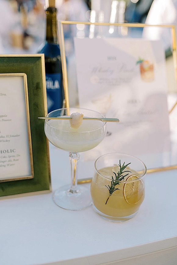 Wedding signature drinks displayed in cocktail glasses beside a signature cocktail bar menu in gold frames on a white bar top, citrus garnish and rosemary sprig