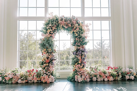 Wedding floral arch with garden rose clusters, hydrangeas and greenery, framed by sunlit grid windows and ground floral arrangements.
