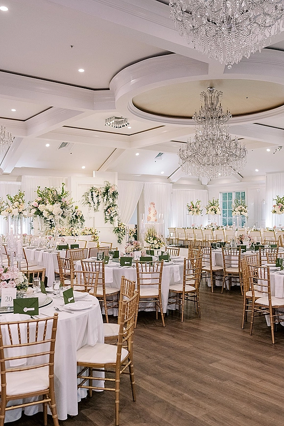 Reception tablescape in a wedding ballroom reception with round tables, white linens, gold chiavari chairs, and tall floral centerpieces under chandeliers