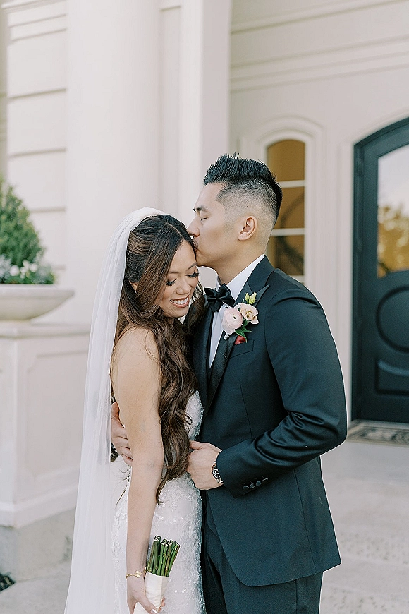Couple portrait of groom kissing bride’s forehead as they embrace, her long veil and lace dress against a light stucco wall backdrop