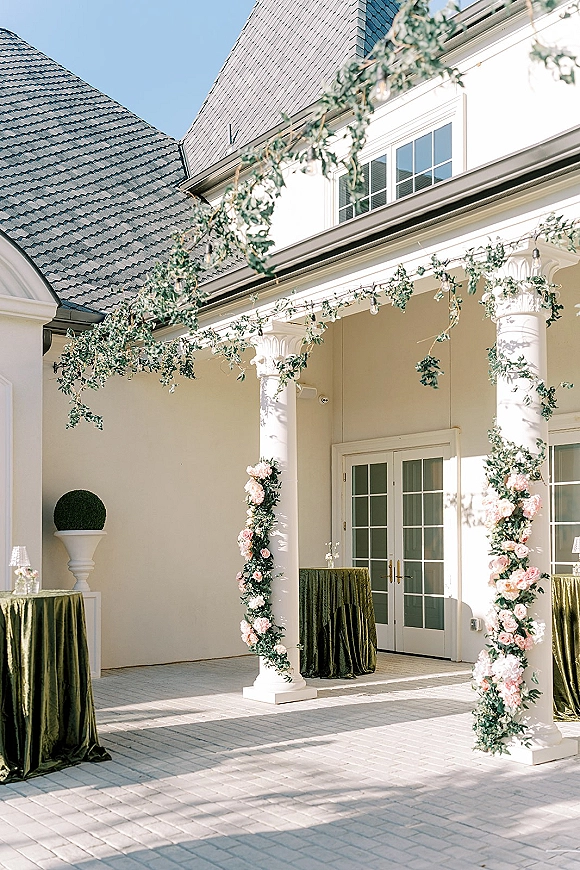 Wedding entrance decor with greenery wedding entrance garland, string lights, and pink rose-wrapped porch columns beside velvet cocktail tables.