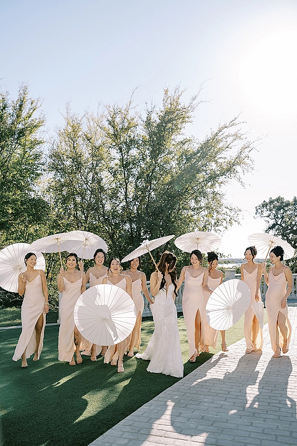 Bride with bridesmaids walking along a garden lawn and brick walkway, bridesmaids with parasols in blush dresses under bright sky