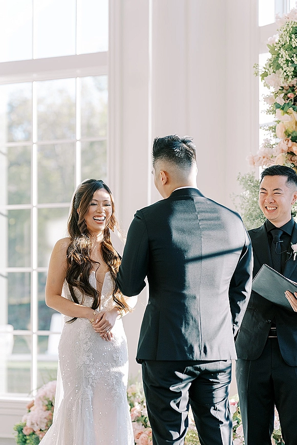 Wedding vows as bride laughs holding groom’s hands beneath a white and blush floral arch in window-lit indoor ceremony space