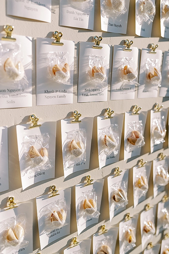 Wedding escort cards on a white display board, each name card clipped to a bagged fortune cookie, with crisp sunlight shadows behind