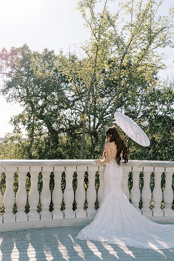 Bridal portrait of a bride holding parasol, showing a lace wedding dress with long train on a sunlit stone balcony terrace with trees behind