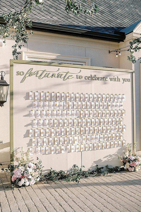 Wedding seating chart in a gold frame with calligraphy escort card display, greenery garland, florals, and string lights against a white wall