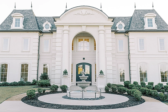 Wedding venue exterior with a front fountain, columned arched entryway, double doors, and trimmed hedges along the walkway on a bright lawn