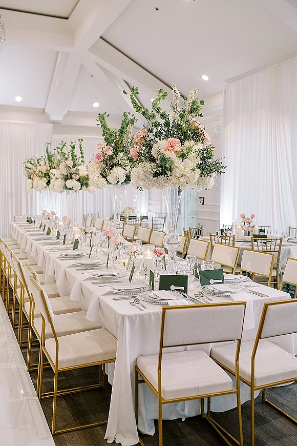 Reception tablescape with long banquet table setup, white linens, gold chairs, tall hydrangea and pink rose centerpieces in a draped hall