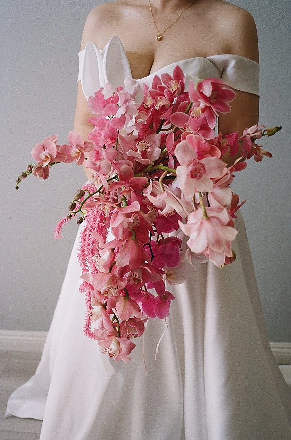 Bridal bouquet of pink orchid bouquet blooms cascading down against a white off-shoulder wedding dress, set before a simple gray wall