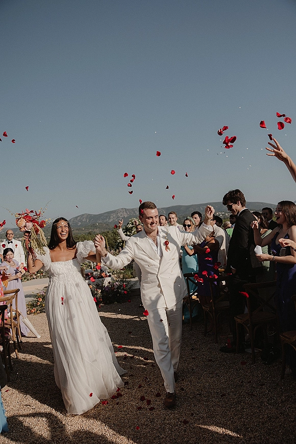 Wedding recessional with rose petal toss as bride in a flowy tulle dress and groom in a white suit walk down a gravel aisle with mountains behind
