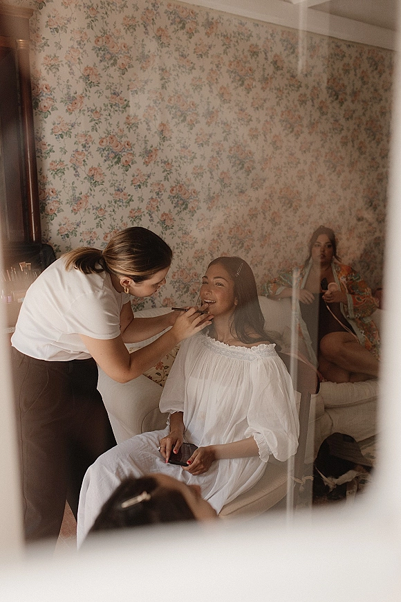Bridal makeup as bride getting ready in a robe with gold hoop earrings, using a brush near floral wallpaper in a bedroom suite
