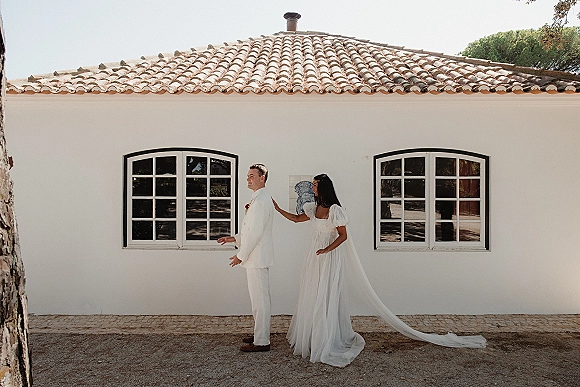 First look moment as bride taps groom’s shoulder, her puff sleeve gown and long veil behind him in a white suit by stucco wall