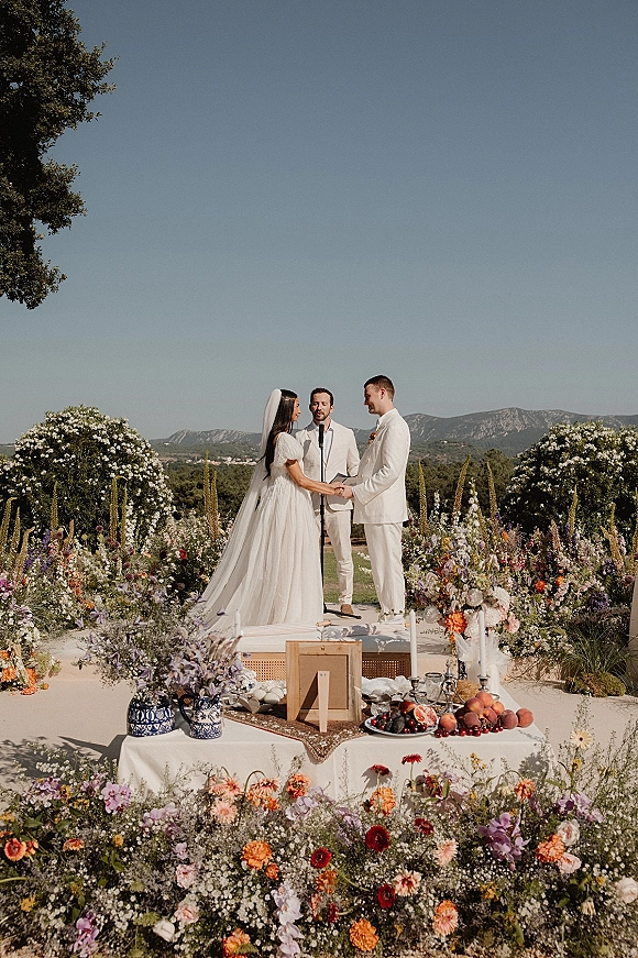 Wedding vows at an outdoor wedding ceremony as the couple holds hands on a floral platform with a veil, microphone, and mountain view backdrop