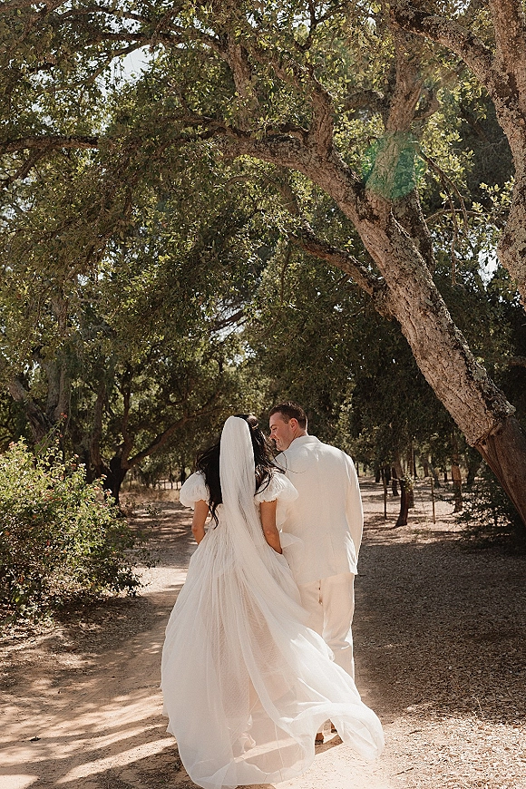 Couple portrait of bride and groom walking away hand in hand, her long veil and dress train flowing on a sunlit wooded path