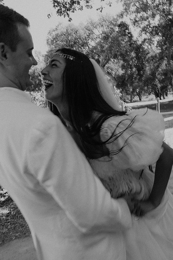 Couple portrait in a black and white wedding photo, bride and groom hugging and laughing on a park lawn with trees and a wooden fence