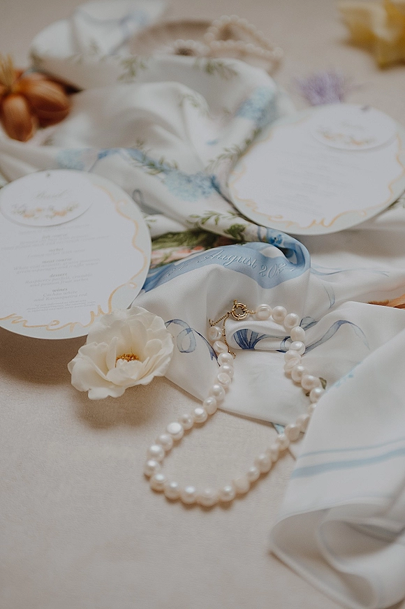 Wedding invitation suite flat lay with letterpress cards, pearl necklace, printed silk scarf, flowers, and twine on linen backdrop