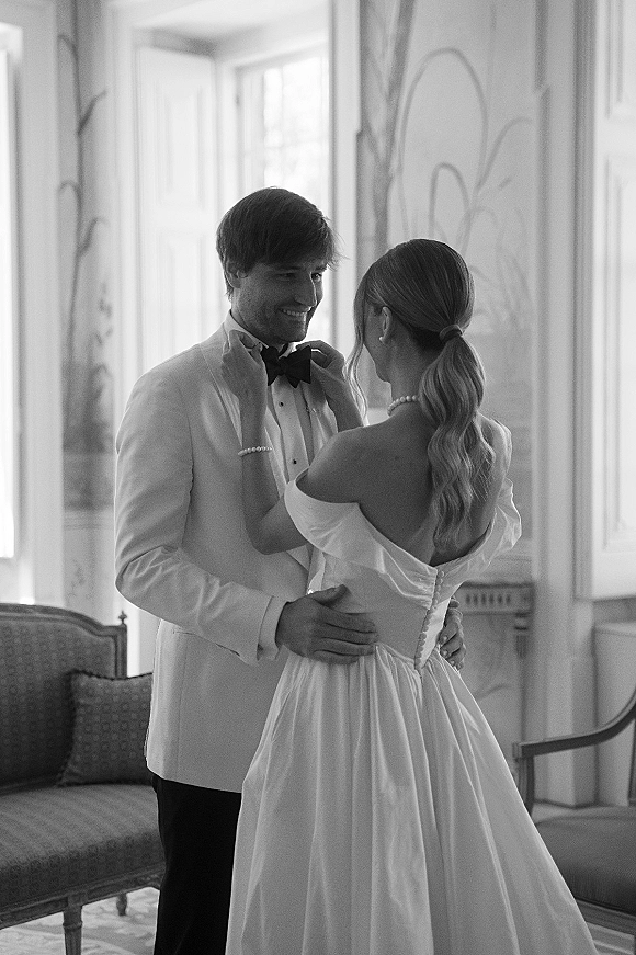 Couple portrait in a vintage room, bride in an off-shoulder gown adjusts groom’s black bow tie by sunlit windows near a sofa