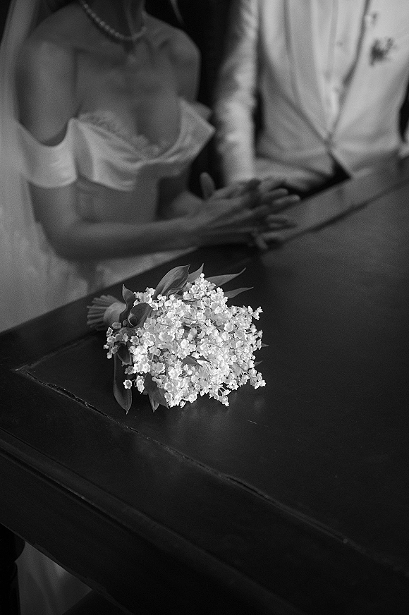 Ceremony moment of bride and groom holding hands, her veil and pearl necklace visible beside a baby's breath bouquet on church pew