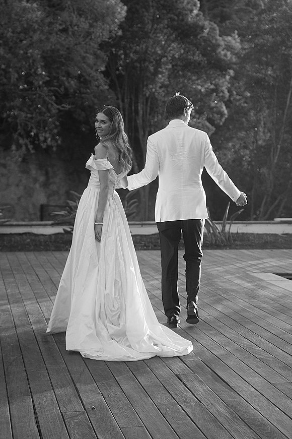 Couple portrait in a black and white wedding portrait, bride and groom holding hands on a sunlit wood deck with garden trees behind