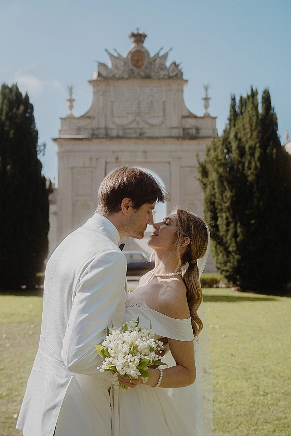 Wedding kiss portrait of bride and groom kissing under an ornate archway, bride in off-shoulder gown with veil holding lily of the valley bouquet