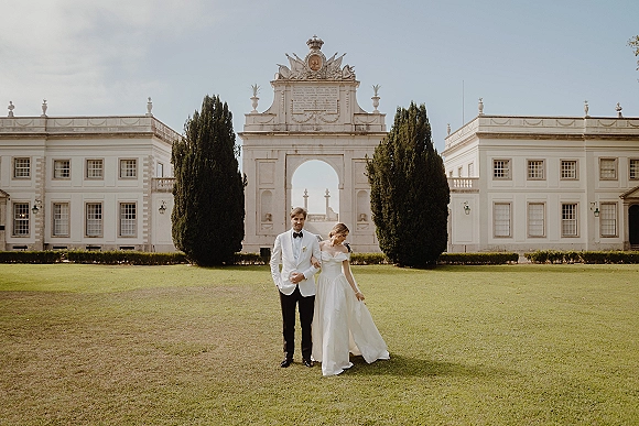 Couple portrait of bride and groom standing together on a garden lawn before a stone archway and grand manor, bride holding dress hem