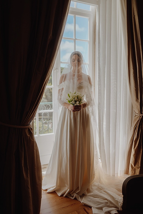 Bridal portrait of a bride holding bouquet with a cathedral veil over her face by a tall window in soft daylight, pearl necklace visible