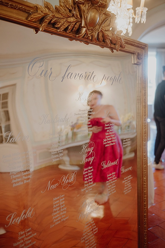 Wedding seating chart mirror in an ornate gold frame with calligraphy lettering, set beneath a chandelier in a white-paneled reception room