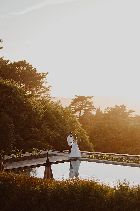 Couple portrait of bride and groom by pool, holding hands at sunset on a terrace with reflecting water, trees, and hills behind