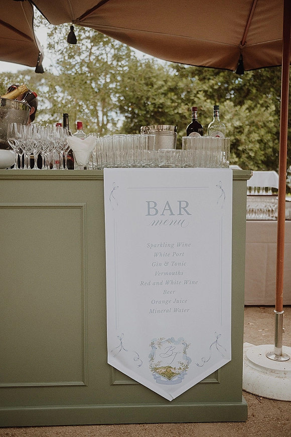 Wedding bar menu sign displayed at an outdoor wedding bar with champagne bottles, liquor, glassware, and a patio umbrella under trees