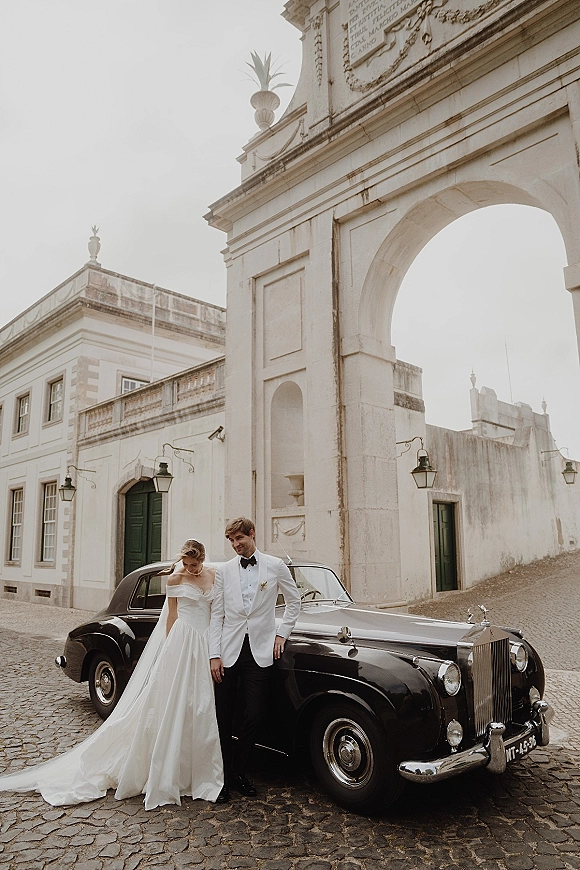 Couple portrait of bride and groom with car, her veil and off-shoulder gown beside a vintage getaway car under a stone archway
