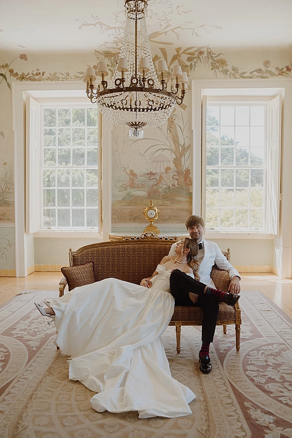 Couple portrait of bride and groom on sofa, her veil draped as she rests on him beneath a chandelier in bright window light
