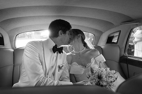 Wedding kiss portrait of newlyweds in car, bride in off-shoulder lace gown and veil kissing groom in white tux in a vintage backseat