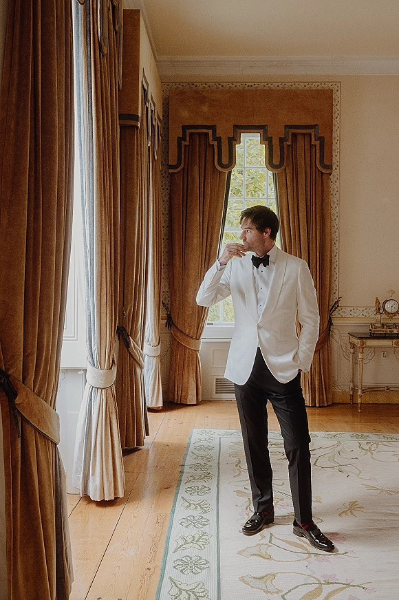 Groom portrait in a white tuxedo jacket and black bow tie, standing by tall windows with velvet drapes in a classic room