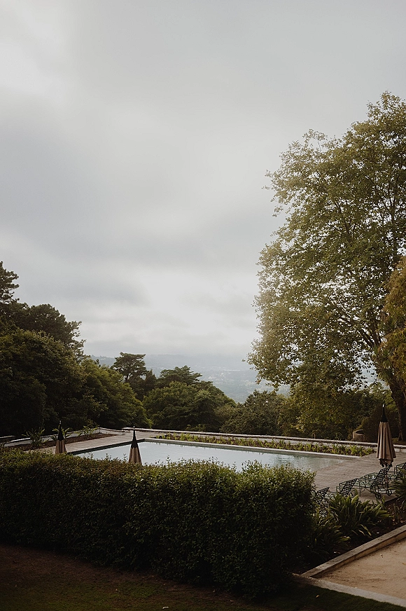 Wedding venue landscape with a pool, patio umbrellas, and lounge chairs bordered by hedges, overlooking a tree-lined hillside under cloudy sky