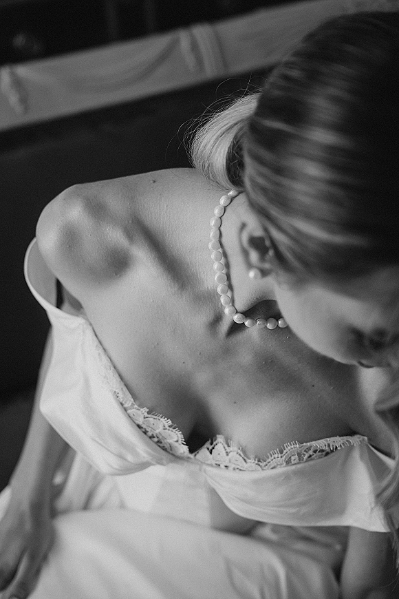 Bridal portrait in black and white of a bride looking down, seated by an upholstered chair, wearing a strapless lace neckline and pearl necklace