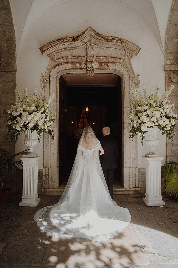 Processional moment as bride enters church with her father, cathedral veil and long dress train framed by white floral urns at stone arch doorway