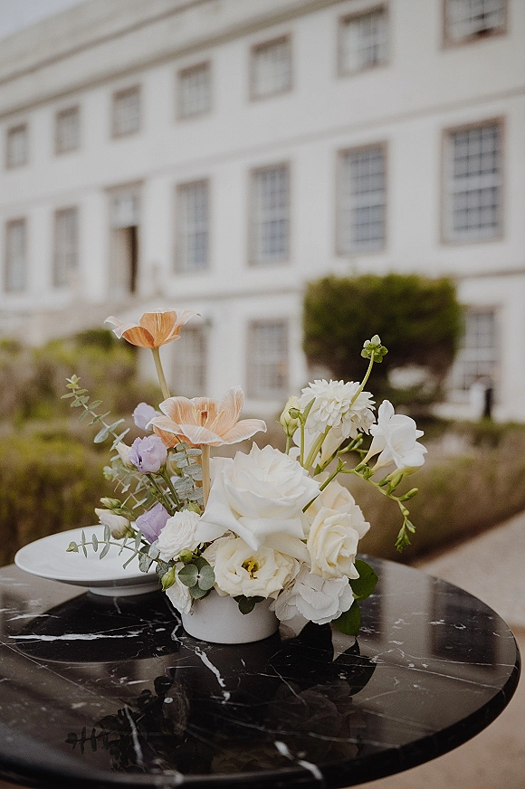 Wedding centerpiece with low floral centerpiece of white roses, peach anemones, dahlias, and eucalyptus in a ceramic vase on marble terrace table