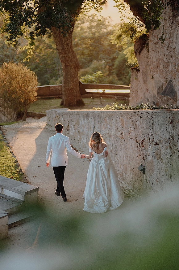 Wedding couple portrait of bride and groom walking away hand in hand, her off-shoulder gown flowing beside his white dinner jacket on a sunset garden path