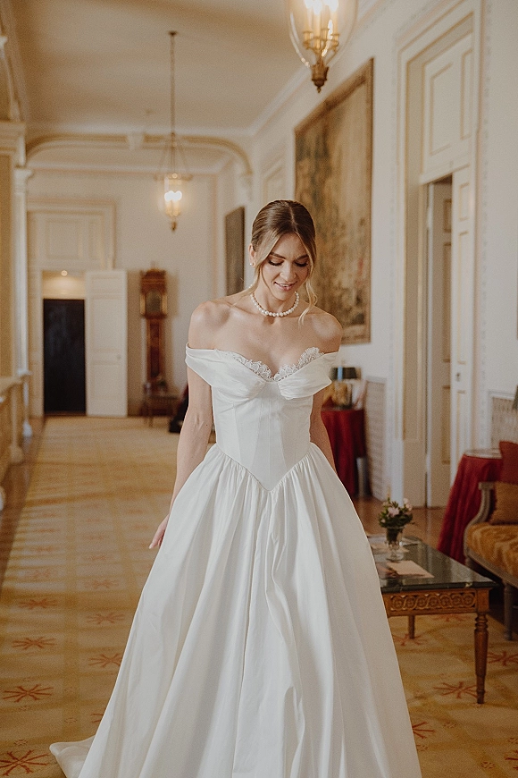 Bridal portrait of a bride in an off shoulder wedding dress with corset bodice and pearl necklace in an ornate hallway with sconces and framed art