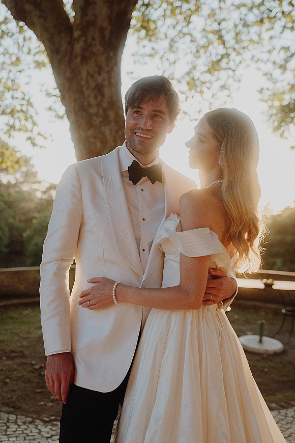 Couple portrait of bride and groom embrace in golden sunset light on an outdoor terrace, her off-shoulder satin gown and pearl jewelry glowing