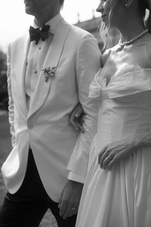 Couple portrait of bride and groom close up, her lace strapless dress with pearl necklace as she holds his arm in sunlit greenery