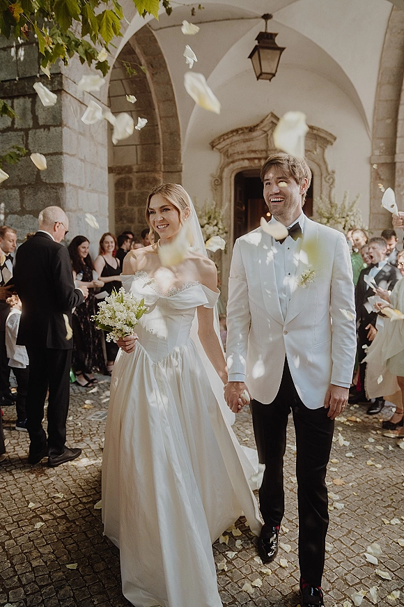 Wedding recessional as bride and groom walk hand in hand out of ceremony under a stone archway, petals flying, bouquet and veil flowing